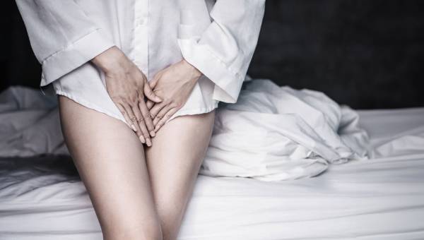 Ein Frau im Nachthemd sitzt auf ihrem Bett und hält schützend die Hände vor ihren Intimbereich